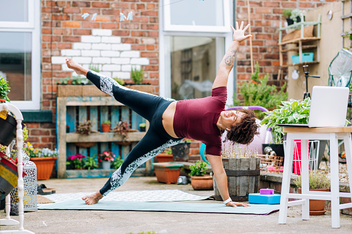A mixed-race mid-adult female yoga instructor performing a Side Star Plank, she is hosting an online class from her back yard on her laptop to her clients during the Corona Virus Pandemic.