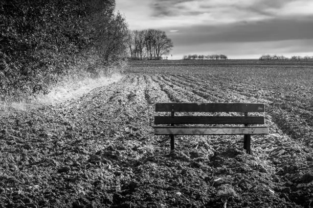 Moody black and white landscape of an old wooden bench in a ploughed field under a cloudy sky in an atmospheric agricultural landscape