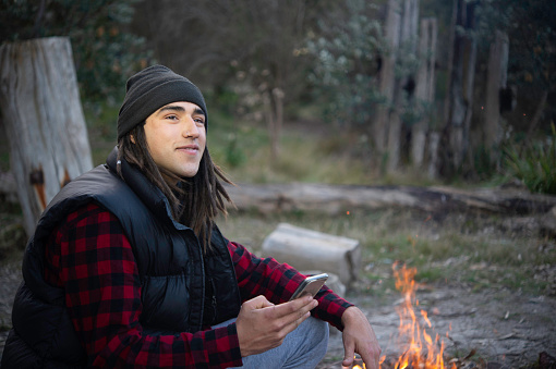 Individual young man having fun outdoors in Australia's natural environment with a couple of dogs, and a fire. Using iPhone for GPS, communication. Aboriginal Australian Ethnicity.