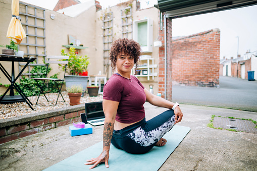 A mixed-race mid-adult female yoga instructor performing a seated spinal twist on an exercise mat. She is hosting an online class from her back yard on her Laptop to her clients during the Corona Virus Pandemic.