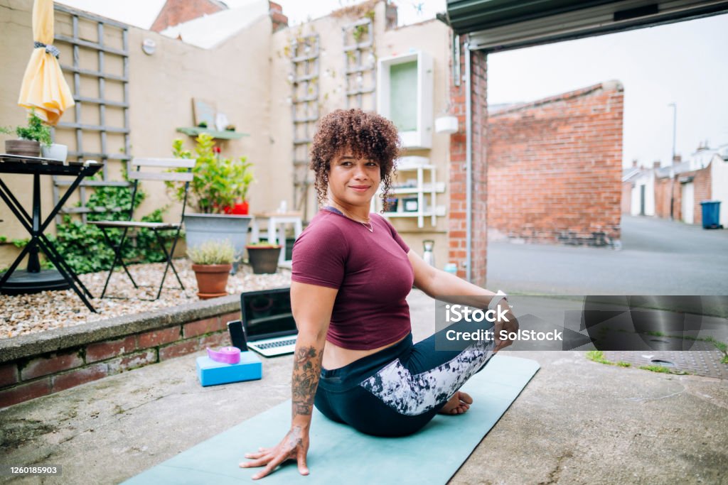 Yoga is Her Passion A mixed-race mid-adult female yoga instructor performing a seated spinal twist on an exercise mat. She is hosting an online class from her back yard on her Laptop to her clients during the Corona Virus Pandemic. Yoga Stock Photo