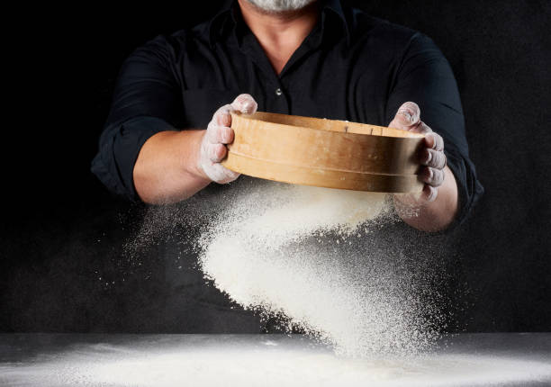 chef a man in a black uniform holds a round wooden sieve in his hands and sifts white wheat flour chef a man in a black uniform holds a round wooden sieve in his hands and sifts white wheat flour on a black background, the particles fly in different directions sifting stock pictures, royalty-free photos & images