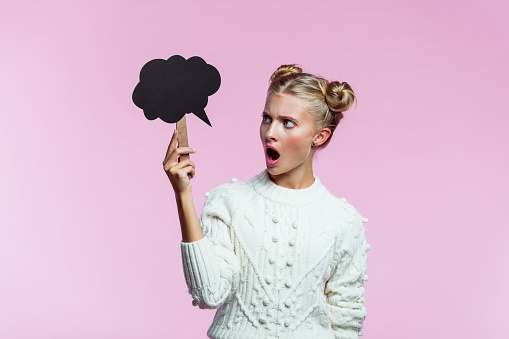 Portrait of angry teenager with hair buns wearing white sweater. Girl holding black speech bubble in hand, looking away and screaming. Studio shot on pink background.