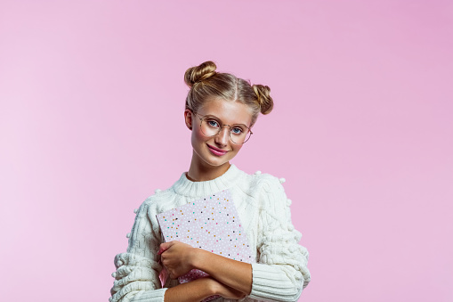 Portrait of happy teenager wearing white sweater and eyeglasses. High schools female student holding pink diary in hands. Studio shot on pink background.