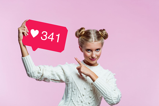 Portrait of happy teenager with hair buns wearing white sweater. Girl holding speech bubble in hand with many hearts, Instagram sign. Studio shot on pink background.