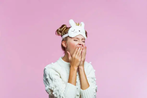 Portrait of teenager wearing white sweater and cat blindfold sleep mask on her forehead, yawning with eyes closed. Studio shot on pink background.