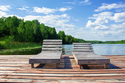 Wooden sun loungers in scandinavian style on wooden pier on lake. Outdoor recreation on sunny summer day. Lake, forest and blue sky. New format vacation. No people, keeping distance.