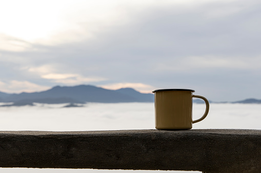 Close up a yellow tin cup of hot coffee with  fog on the mountain background in the morning.