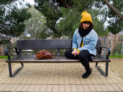 Young adult woman with yellow woolen cap sitting on a park bench thoughtful. Autumn urban concept.