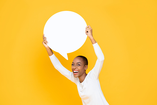Cheerful happy young African American woman smiling and holding speech bubble with empty space for text overhead isolated on yellow background