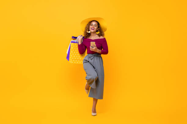 happy african american woman carrying shopping bags and phone raising one leg - women beautiful studio shot full length imagens e fotografias de stock