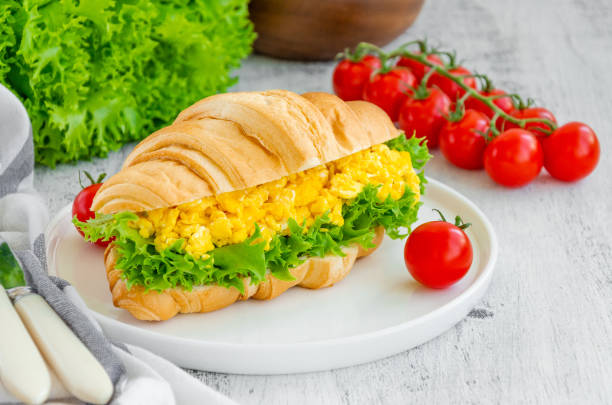istock Croissant with scrambled eggs, lettuce and cherry tomatoes on a white plate with ingredients to cook on a wooden background. Horizontal, copy space. 1260485569