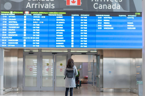Welcome home! A young female is standing in front of the arrival board waiting to be reunited with her husband.  She is wearing a protective face mask. Travelling during the COVID-19 pandemic. airports canada stock pictures, royalty-free photos & images