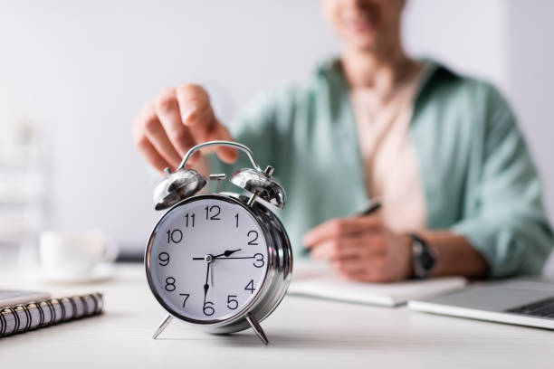 istock Selective focus of man pulling hand to alarm clock near laptop and notebooks on table, concept of time management 1260568303