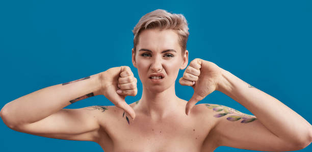 portrait of a young attractive half naked tattooed woman with perfect skin looking dissatisfied, showing thumbs down isolated over blue background - women tattoo naked sadness imagens e fotografias de stock