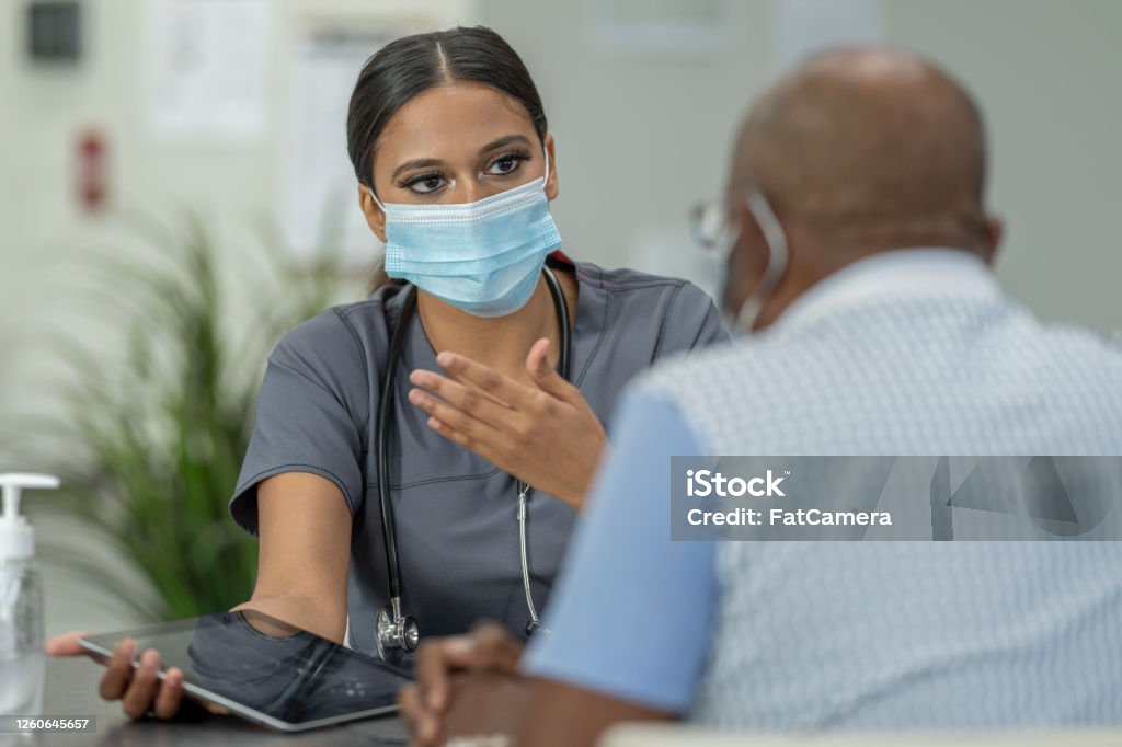 Masked Medical Appointment A senior patient and a female doctor meet in a medical clinic while wearing protective face masks to avoid the transfer of germs. Patient Stock Photo