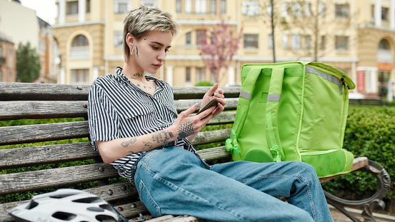 Young delivery woman with thermo bag or backpack checking order using smartphone, sitting on the bench outdoors. Courier, delivery service concept. Focus on woman. Web Banner