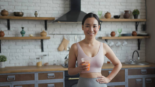 zdrowa azjatka stojąca w kuchni z pomarańczowym świeżym sokiem w rękach - juice glass healthy eating healthy lifestyle zdjęcia i obrazy z banku zdjęć