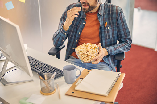 Close up of male worker enjoying crunchy corn snack while sitting at the table with computer, documents and cup of coffee