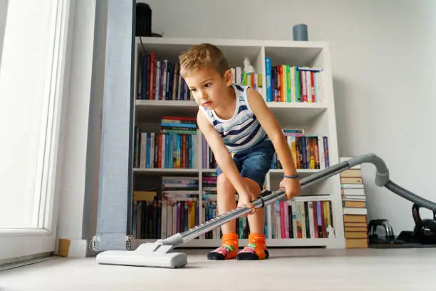 Photo of Little kid using vacuum cleaner at home - Small boy cleaning floor in apartment - Child doing housework having fun - side view full length in summer day - childhood development real people concept