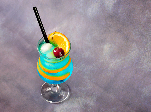 A glass of blue lagoon cocktail with a cherry, a slice and peel of an orange and ice stands on a gray background. Close-up.