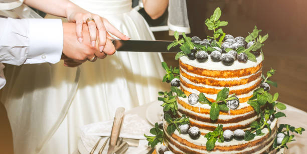 istock Wedding couple is cutting modern rustic cake. Open sponge dessert with mint leaves and fresh fruit grapes on top. Boho style wedding cake. Groom in the black suit and bride in white elegant dress. 1261118898