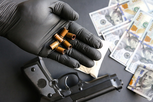 Gun lying on the table. Man in black gloves holding bullets. Illegal drug selling. Criminal problems. Dollars.