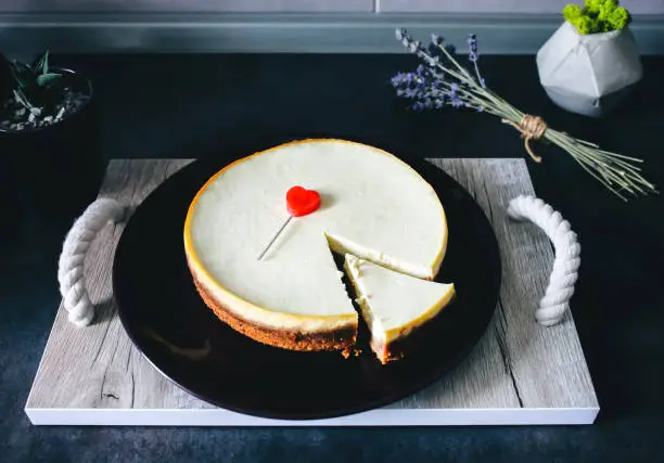 Classical homemade New York cheesecake is lying on dark-violet plate on wooden tray with rope handles on kitchen grey table. Lavender and succulent  on background. Red heart to Valentine's day present