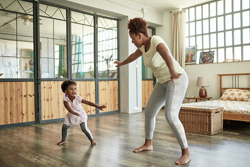 Afro-Caribbean mother in mid 20s and 3 year old daughter having fun doing dance exercises at home in apartment bedroom.