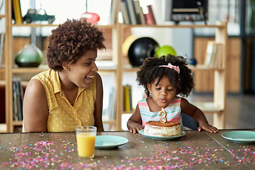 Partial front view of Afro-Caribbean single mother in mid 20s laughing as she watches daughter blow out candle on birthday cake.