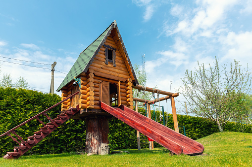 Small wood log playhouse hut with stairs ladder and wooden slide on children playground at park or house yard. Green grass lawn garden and blue clear sky in background on bright sunny summer day.