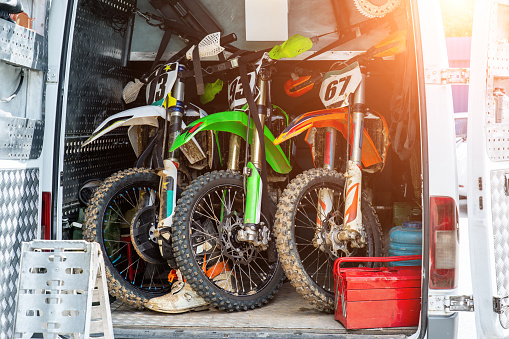 Three bright dirty sport motocross motorcycles stand in tail of technical support assistance van in team camp during extreme offroad championship. Mobile cycle service, maintenance transport concept.