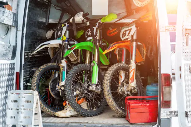 Three bright dirty sport motocross motorcycles stand in tail of technical support assistance van in team camp during extreme offroad championship. Mobile cycle service, maintenance transport concept.