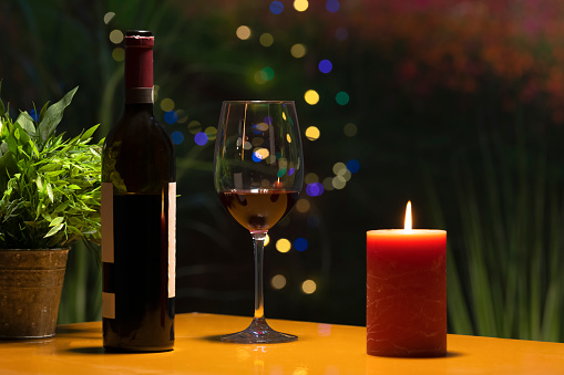 Close up of a glass of wine surrounded by a wine bottle and a lit candle on a table on an out of focus background. Celebration and holidays concept.