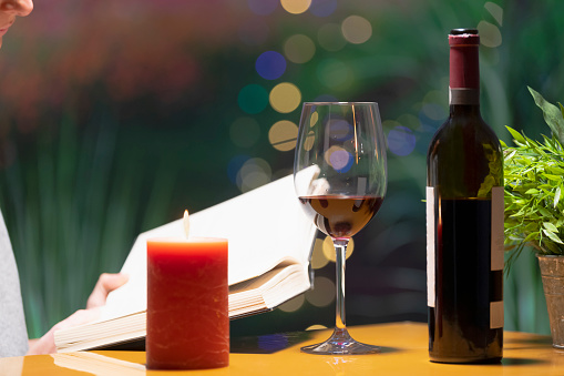 Close up of a glass of wine surrounded by a lit candle and an open bottle of wine, with an open book being held by a person behind on an out of focus background. Leisure and lifestyle concept.