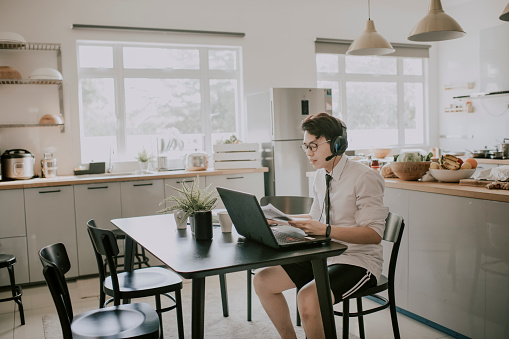 Un joven adulto chino asiático con corbata, camisa de manga larga y pantalón corto sentado usando portátil en su comedor para videoconferencia con su socio de negocios y colega reunión de negocios virtual photo