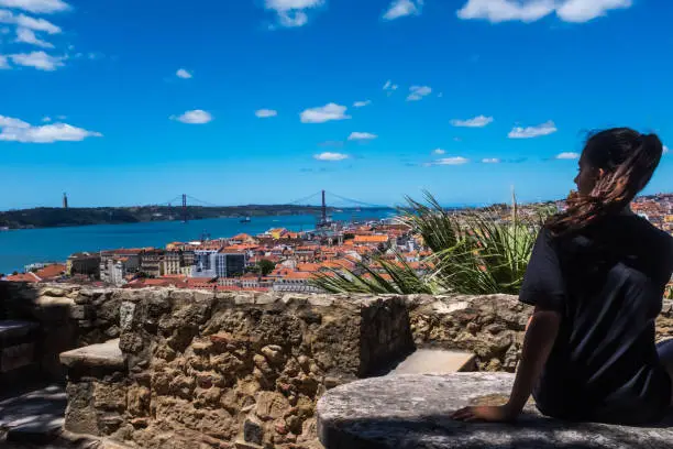 Profile view of a teenager overlooking the urban landscape of Lisbon with its red roofs and the Ponte 25 de Abril (25th of April Bridge) in the background. Bright and windy day, the typical weather conditions in Portugal.