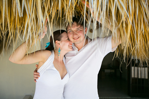 couple in love dressed in white, stands under a roof of palm leaves.
