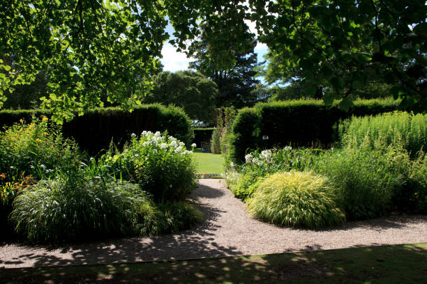 A serene garden scene shows a meticulously maintained landscape featuring colorful blooms surrounded by verdant grass and large trees, creating a calm and idyllic springtime atmosphere.