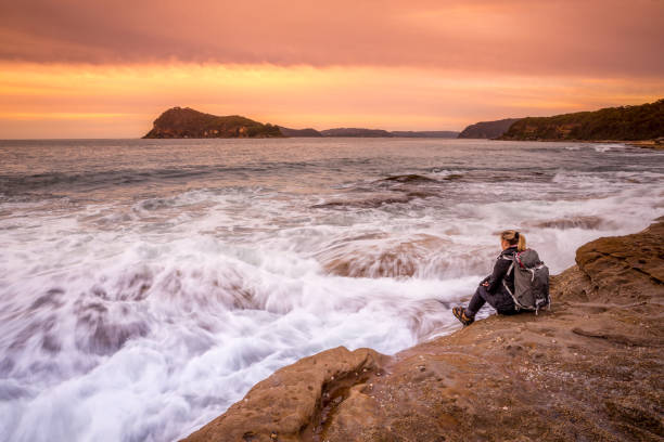 istock Woman sitting on rocks watching the waves crash over the rocks 1261394721