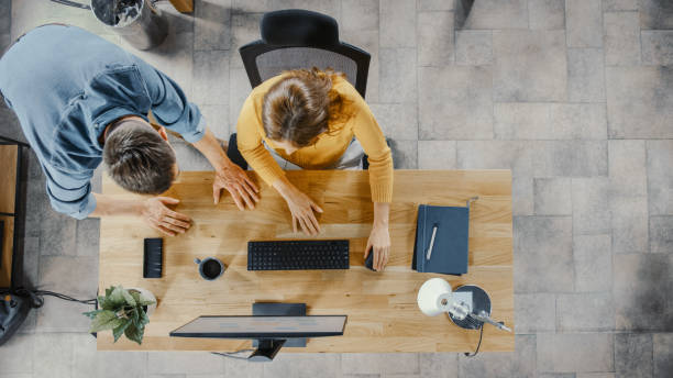 istock Top View Shot: Female Office Worker Uses Desktop Computer, Project Manager Stands Beside and gives Advice on Optimizing Workflow for Customer Experience Management. 1261396380