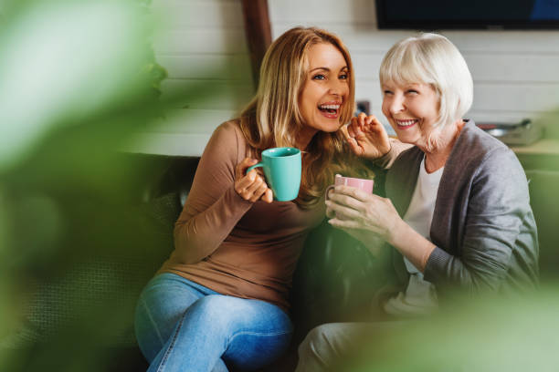 mãe mais velha feliz com filha adulta sentada no sofá e segurando xícaras com café ou chá em casa. conceito de união - family retirement smiling multi generation family - fotografias e filmes do acervo