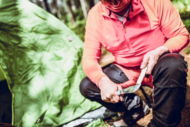 Senior Male Sharpening Knife While Sitting Near Tent In Forest