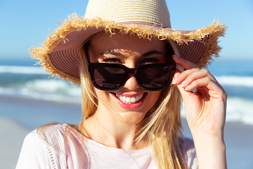 Portrait of attractive blonde Caucasian woman enjoying time at the beach on a sunny day, looking at camera and smiling, with blue sky and sea in the background.