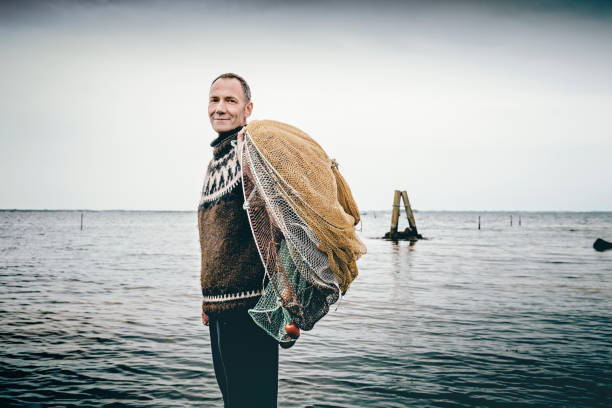 Candid portrait of a shrimp fisherman preparing his nets in the shallow water. Colour, horizontal with some copy space. The net is left in the water for 24 hours before it is hauled in and the catch sorted. Photographed on the island of Nyord in Denmark. He is fishing for salt water shrimp in the local fjord’s and they are highly sought after as a delicacy here in Denmark.