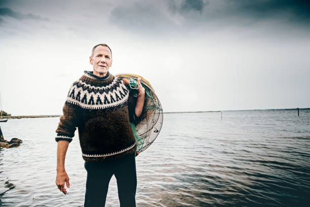 Candid portrait of a shrimp fisherman preparing his nets in the shallow water. Colour, horizontal with some copy space. The net is left in the water for 24 hours before it is hauled in and the catch sorted. Photographed on the island of Nyord in Denmark. He is fishing for salt water shrimp in the local fjord’s and they are highly sought after as a delicacy here in Denmark.