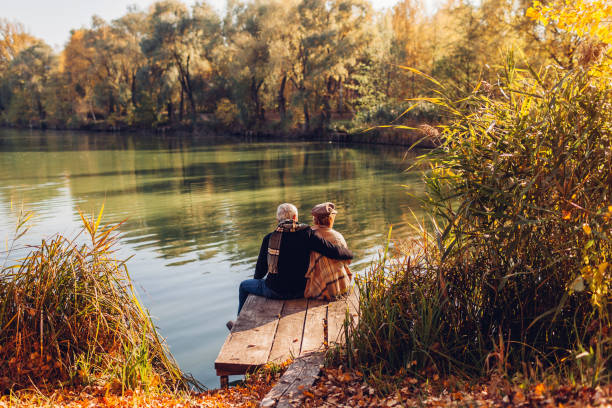 senior family couple relaxing by autumn lake. man and woman enjoying nature and hugging sitting on pier - forest sitting men comfortable imagens e fotografias de stock