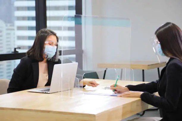 Manager from HR department wearing facial mask is interviewing new applicant who is handing her resume and profile through the partition for social distancing and new normal policy