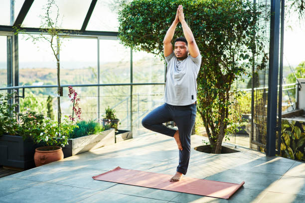 Young man practicing the tree pose in a yoga studio Young man in sportswear practicing the tree pose on an exercise mat during a yoga session in a bright modern studio good posture stock pictures, royalty-free photos & images