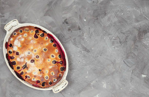 Portion of peach clafoutis, an eggy custard baked with sweet summer fruit, a creamy texture with sliced peaches and icing sugar dressing in a plate, the most cherished home-baked desserts in France made with fresh peaches, vanilla extract,  sugar, lemon zest, eggs, milk, heavy cream, flour, ground cardamom, salt and dusted with icing sugar